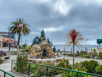 A coastal scene featuring a large sculpture with multiple figures atop a rocky structure. Surrounding the sculpture are well-manicured gardens with various plants and flowers. An American flag waves beside a tall palm tree on the left. Nearby, there are some buildings, including one labeled 'Fish Hopper'. The ocean is visible in the background under a cloudy sky.