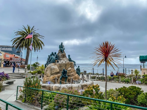 A coastal scene featuring a large sculpture with multiple figures atop a rocky structure. Surrounding the sculpture are well-manicured gardens with various plants and flowers. An American flag waves beside a tall palm tree on the left. Nearby, there are some buildings, including one labeled 'Fish Hopper'. The ocean is visible in the background under a cloudy sky.