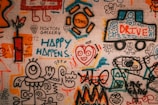 A vibrant graffiti mural covers a brick wall, featuring abstract and cartoonish figures, playful scribbles, and colorful text. Various elements such as a truck labeled 'Drive,' a happy face with a heart, and phrases like 'Happy Happens' and 'Blow Up the Rules' are visible. The artwork is filled with energetic and whimsical shapes, blending together in a collage of creativity.