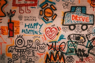 A vibrant graffiti mural covers a brick wall, featuring abstract and cartoonish figures, playful scribbles, and colorful text. Various elements such as a truck labeled 'Drive,' a happy face with a heart, and phrases like 'Happy Happens' and 'Blow Up the Rules' are visible. The artwork is filled with energetic and whimsical shapes, blending together in a collage of creativity.