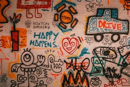 A vibrant graffiti mural covers a brick wall, featuring abstract and cartoonish figures, playful scribbles, and colorful text. Various elements such as a truck labeled 'Drive,' a happy face with a heart, and phrases like 'Happy Happens' and 'Blow Up the Rules' are visible. The artwork is filled with energetic and whimsical shapes, blending together in a collage of creativity.