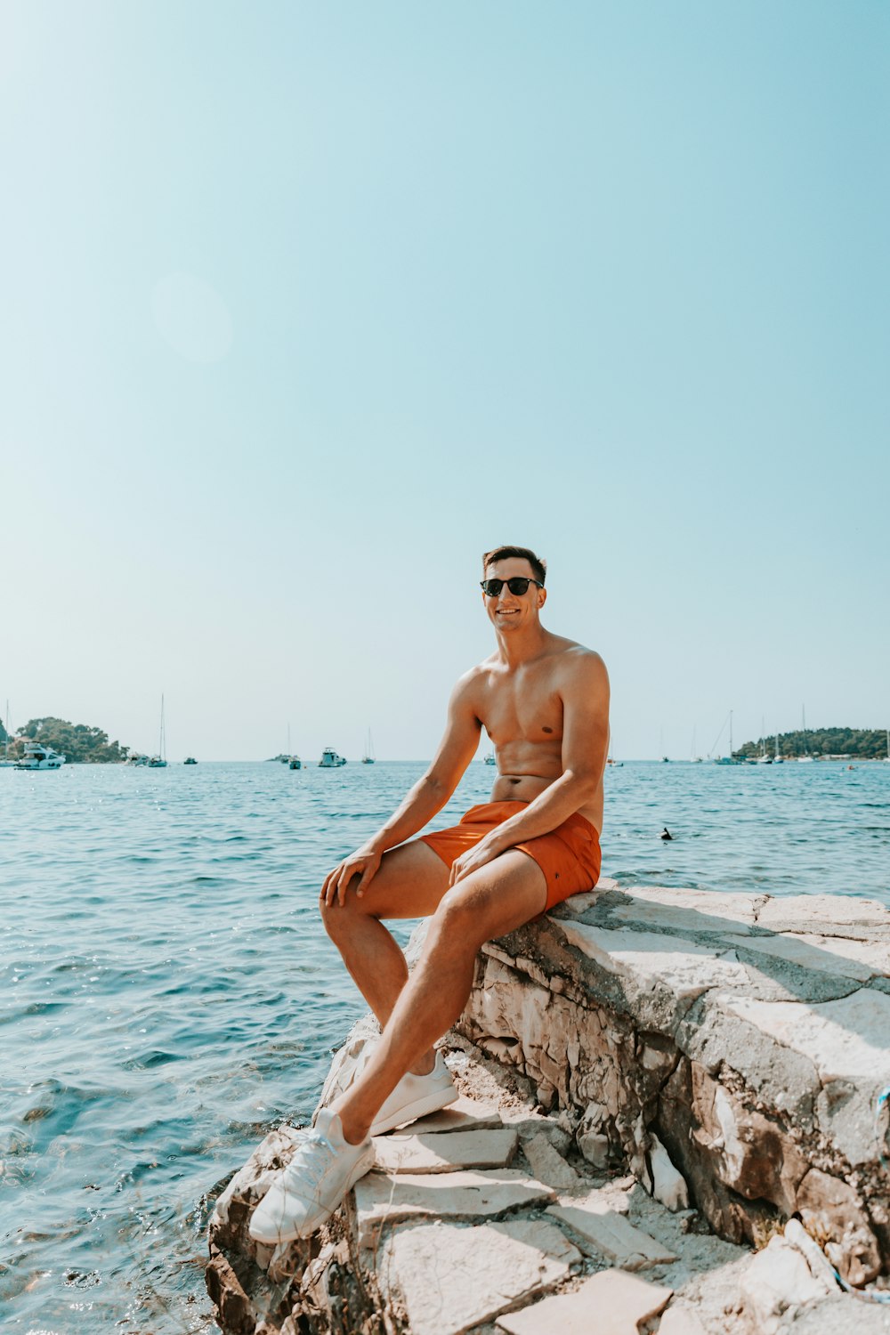 topless man in red shorts sitting on rock near body of water during daytime