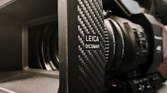 Close-up of a professional video camera lens with the branding 'LEICA DICOMAR' visible on the side. The lens is housed in a textured, dark casing, and intricate details of the camera's focus ring and controls are apparent.