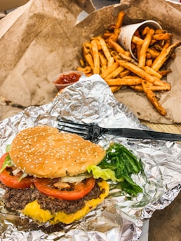A cheeseburger with lettuce, tomato, and a sesame seed bun sits on a piece of aluminum foil. Next to it is a serving of French fries in a paper cup, spilling onto a paper surface. Ketchup in a small container is visible nearby.
