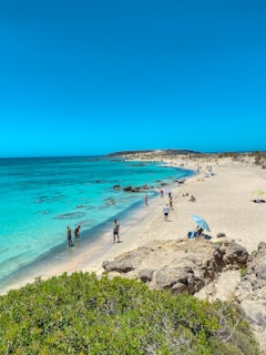 A vibrant beach scene with turquoise water and sunbathers enjoying the sand under clear skies.