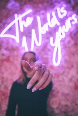 A neon sign with a motivational message glows in purple against a floral-patterned wall. A person in the foreground extends their hand towards the camera, creating a shallow depth of field effect.