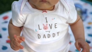 A close-up of a baby wearing a white onesie with the text 'I Love my Daddy' and a cute pink bunny design. The baby's skin tone is visible, and their hands are positioned near the text. A multi-colored blanket with abstract patterns is partially visible in the background.