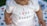 A close-up of a baby wearing a white onesie with the text 'I Love my Daddy' and a cute pink bunny design. The baby's skin tone is visible, and their hands are positioned near the text. A multi-colored blanket with abstract patterns is partially visible in the background.