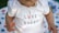 A close-up of a baby wearing a white onesie with the text 'I Love my Daddy' and a cute pink bunny design. The baby's skin tone is visible, and their hands are positioned near the text. A multi-colored blanket with abstract patterns is partially visible in the background.