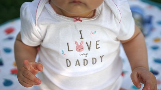 A close-up of a baby wearing a white onesie with the text 'I Love my Daddy' and a cute pink bunny design. The baby's skin tone is visible, and their hands are positioned near the text. A multi-colored blanket with abstract patterns is partially visible in the background.