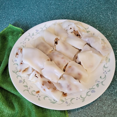 A plate of steamed rice rolls filled with a mixture of minced meat and possibly vegetables, arranged neatly. The rolls have a smooth, translucent appearance and are placed on a decorative white plate with floral patterns. A green textured cloth is beside the plate, set on a speckled green surface.