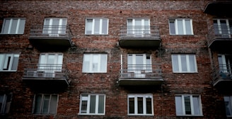 A brick apartment building facade with several windows and balconies. The structure appears aged but sturdy, with simple metallic railings on each balcony. The overall architectural style is utilitarian.