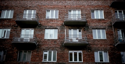 A brick apartment building facade with several windows and balconies. The structure appears aged but sturdy, with simple metallic railings on each balcony. The overall architectural style is utilitarian.
