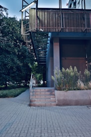 An urban outdoor scene featuring a modern building with a stairway leading up to a platform. The structure is partially surrounded by green foliage and plants growing alongside the building. The architecture is characterized by clean lines and a metallic railing.