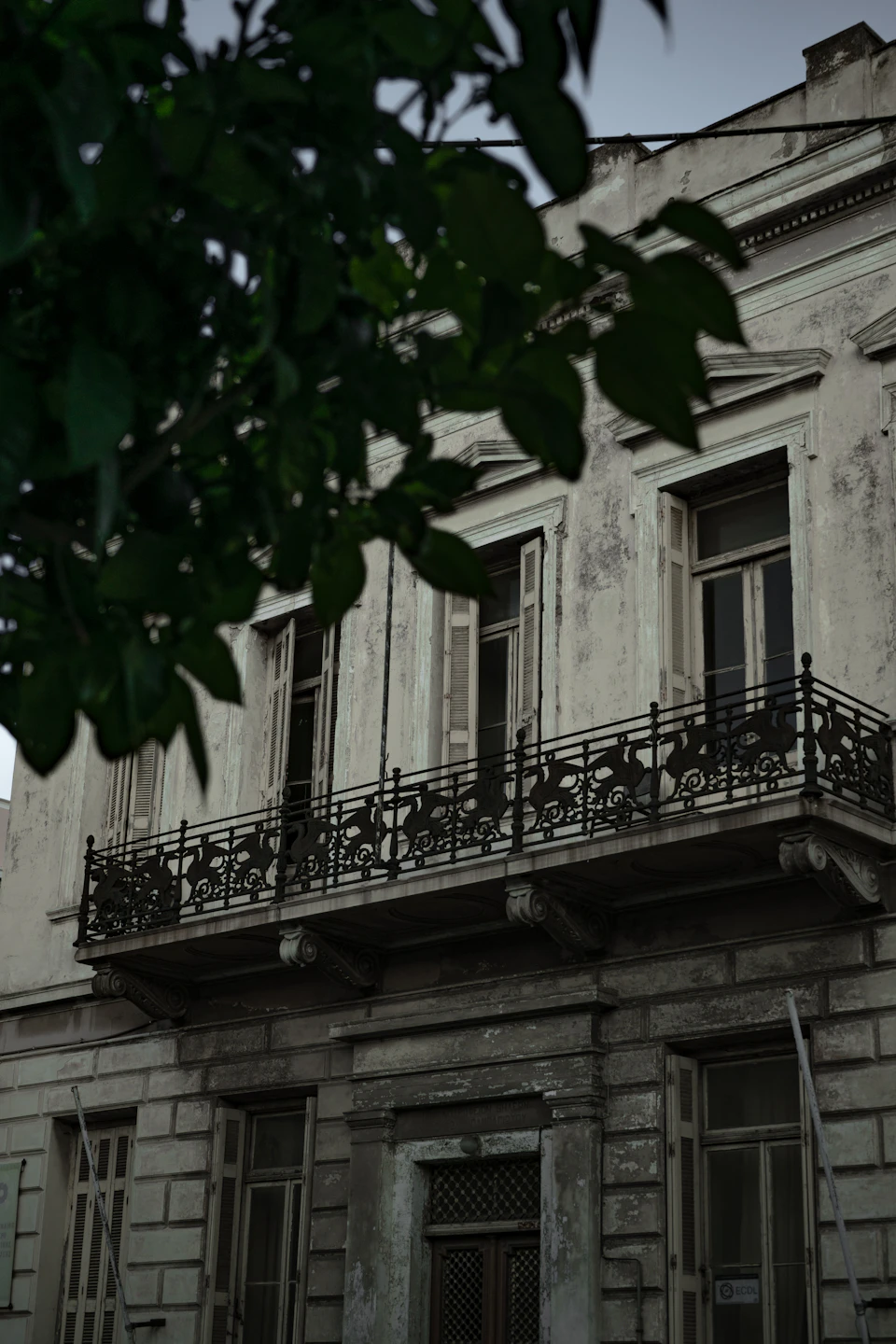 The image depicts a two-story white building with black wrought iron railings on its windows and balcony, situated on a street corner. The building's facade is adorned with green leaves that add a touch of nature to the urban setting. A tree can be seen in front of the building, providing some shade for passersby.