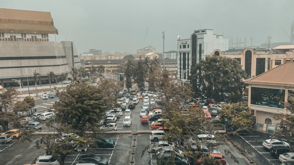 A congested parking lot filled with various cars is surrounded by tall trees and multiple modern buildings in a city setting. The sky is overcast, creating a muted atmosphere.