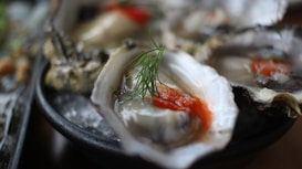 Fresh oysters presented on a bed of ice, garnished with a sprig of dill and a vibrant red sauce, likely cocktail sauce. The image focuses on the detail and texture of the shell and the glossy appearance of the oyster inside.