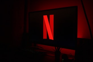 A computer monitor displays a large red 'N' central on a black screen, surrounded by a dark room with a dim red glow. A keyboard and small plant sit below the monitor.
