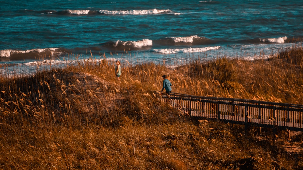 man in black jacket standing on brown wooden fence near sea during daytime