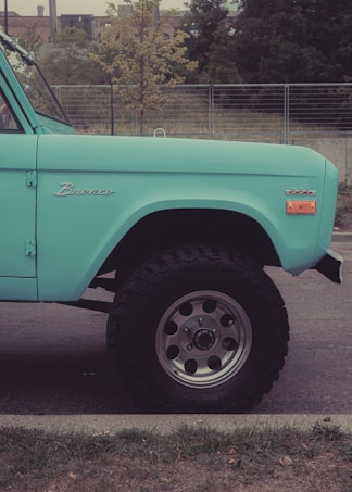 A vintage turquoise vehicle with the name 'Bronco' displayed on its side. The vehicle features large, rugged tires and is parked on a street with a grassy edge. In the background, there are metal railings, trees, and an industrial building.