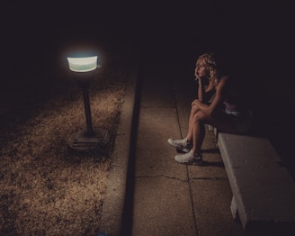 The grey man lost in thought, seated on a worn bench beneath flickering street lamps.