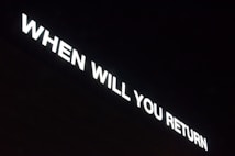 Bright white text reads 'WHEN WILL YOU RETURN' against a dark background, seemingly forming part of a sign or installation.
