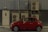 A compact red car is parked beside a building with multiple air conditioning units mounted on a corrugated metal wall. A small door with a rectangular window is visible on the left side of the image.