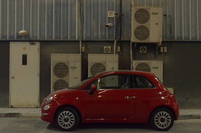 A compact red car is parked beside a building with multiple air conditioning units mounted on a corrugated metal wall. A small door with a rectangular window is visible on the left side of the image.