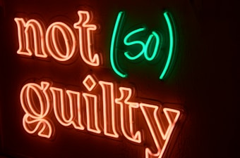 Neon lights display the words 'not (so) guilty' with a black background. The words 'not' and 'guilty' are in orange neon, while '(so)' is in green neon.