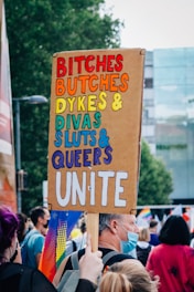A crowd of people gathered outdoors, likely at a rally or pride event. A person holds a large sign with colorful text that reads 'Bitches, Butches, Dykes, Divas, Sluts, Queers Unite.' People in the background are wearing face masks and holding rainbow flags.