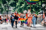 A group of diverse people are marching on a street, some carrying rainbow flags and signs. Many are wearing face masks, and the event seems festive and inclusive, taking place in a sunny outdoor setting with trees and buildings in the background.