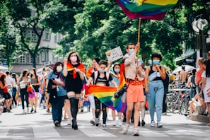 A group of diverse people are marching on a street, some carrying rainbow flags and signs. Many are wearing face masks, and the event seems festive and inclusive, taking place in a sunny outdoor setting with trees and buildings in the background.