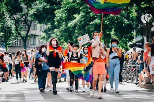 A group of diverse people are marching on a street, some carrying rainbow flags and signs. Many are wearing face masks, and the event seems festive and inclusive, taking place in a sunny outdoor setting with trees and buildings in the background.