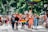 A group of diverse people are marching on a street, some carrying rainbow flags and signs. Many are wearing face masks, and the event seems festive and inclusive, taking place in a sunny outdoor setting with trees and buildings in the background.