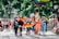 A group of diverse people are marching on a street, some carrying rainbow flags and signs. Many are wearing face masks, and the event seems festive and inclusive, taking place in a sunny outdoor setting with trees and buildings in the background.