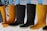 A row of rubber boots, primarily in black and yellow colors, are hanging upside down. The background features a few green leaves and a plain, light backdrop.