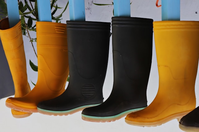 A row of rubber boots, primarily in black and yellow colors, are hanging upside down. The background features a few green leaves and a plain, light backdrop.