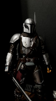 A figure wearing intricately detailed armor with a metallic finish stands in a dark setting. The armor is sleek and includes a helmet with a distinctive visor. The figure holds a weapon and has a belt with various tools and accessories attached.