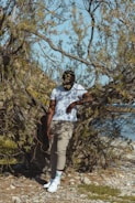 A person stands casually in a natural setting, surrounded by dry, leafless branches and small rocks on the ground. The person is wearing a white t-shirt, beige pants, white sneakers, and a black cap. Sunlight casts shadows on the landscape, and part of a body of water is visible in the background.