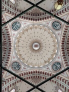 An ornate and intricately designed dome ceiling featuring detailed patterns and calligraphy. The central area of the dome has a symmetrical, circular design with floral motifs, surrounded by alternating sections of red and white arches. Decorative calligraphic elements are positioned at intervals around the dome's interior.