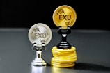 Two coins labeled 'EXU' are standing upright on holders. One is silver and placed on a silver stand, the other is gold and placed on a stack of gold coins. The coins feature leaf designs.