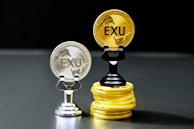 Two coins labeled 'EXU' are standing upright on holders. One is silver and placed on a silver stand, the other is gold and placed on a stack of gold coins. The coins feature leaf designs.