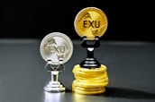 Two coins labeled 'EXU' are standing upright on holders. One is silver and placed on a silver stand, the other is gold and placed on a stack of gold coins. The coins feature leaf designs.