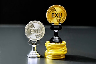 Two coins labeled 'EXU' are standing upright on holders. One is silver and placed on a silver stand, the other is gold and placed on a stack of gold coins. The coins feature leaf designs.