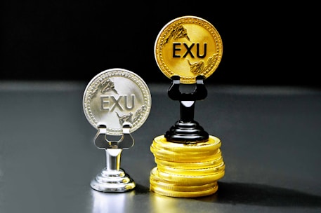 Two coins labeled 'EXU' are standing upright on holders. One is silver and placed on a silver stand, the other is gold and placed on a stack of gold coins. The coins feature leaf designs.
