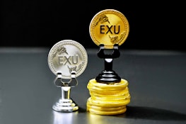 Two coins labeled 'EXU' are standing upright on holders. One is silver and placed on a silver stand, the other is gold and placed on a stack of gold coins. The coins feature leaf designs.