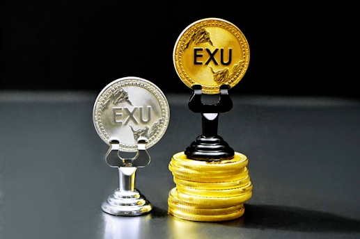 Two coins labeled 'EXU' are standing upright on holders. One is silver and placed on a silver stand, the other is gold and placed on a stack of gold coins. The coins feature leaf designs.