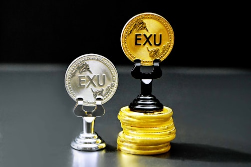 Two coins labeled 'EXU' are standing upright on holders. One is silver and placed on a silver stand, the other is gold and placed on a stack of gold coins. The coins feature leaf designs.