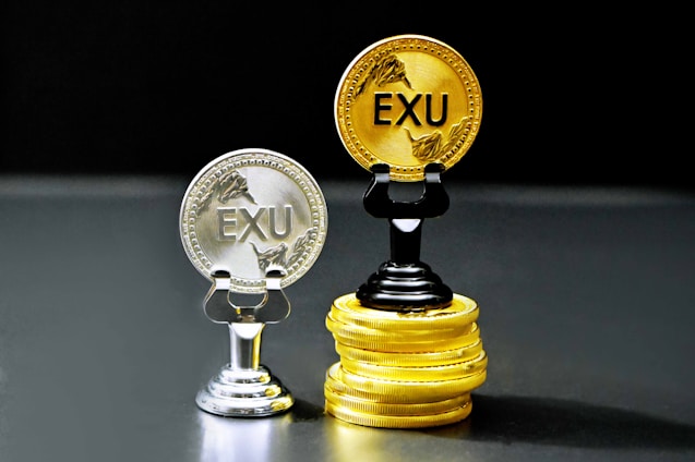 Two coins labeled 'EXU' are standing upright on holders. One is silver and placed on a silver stand, the other is gold and placed on a stack of gold coins. The coins feature leaf designs.