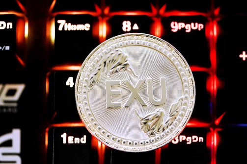 A silver coin with the text 'EXU' embossed in the center, set against an illuminated background of keyboard keys with red and orange lighting. The coin features intricate details like abstract designs around the edges.